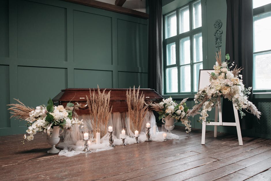 A floral arrangement of white roses, lilies, and orchids, accented with beige pampas grass and green foliage, positioned on a white stand beside a wooden piano draped with soft white fabric. The arran