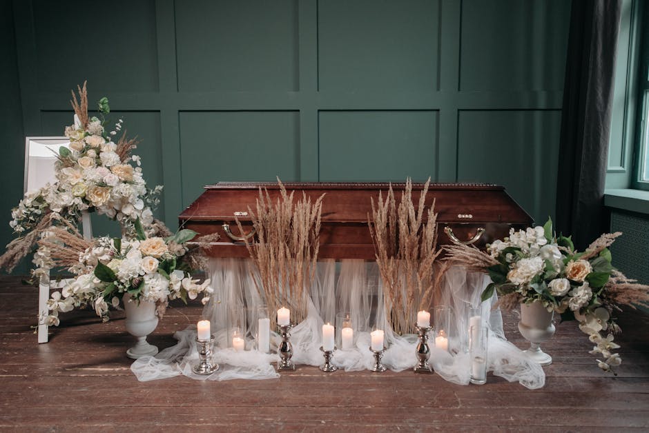A solemn floral arrangement placed on a polished wooden surface in front of a vintage wooden memorial box, featuring white roses, peonies, and eucalyptus leaves, complemented by tall dried pampas gras