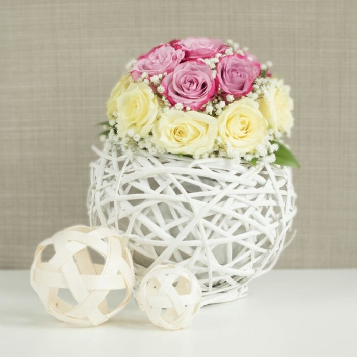 An artfully composed floral centerpiece takes pride of place in this image, the kind of arrangement one might find gracing a light-filled dining room near Grove Park station or a small wedding venue tucked off Burlington Lane. The eye is immediately drawn to the domed bouquet of roses, gathered compactly to create a rounded, almost sculptural form. About a dozen roses in total are used, with colours thoughtfully balanced: soft blush and pastel pinks with a hint of lavender sit alongside creamy butter-yellow and smooth ivory blooms. The petals of each rose appear velvety and fresh, some still curled tightly in bud, others unfolding into elegant spirals that catch the soft light. Threaded through the roses are dainty sprays of white baby's breath, their tiny blossoms resembling scattered stars, adding a light, airy veil and increasing the sense of volume without overwhelming the design. Cradling this bouquet is a distinctive spherical vase fashioned from narrow, twig-like branches or wicker, painted a bright, clean white. These strands weave together in an open, lattice-like pattern, forming a globe that exposes hints of the inner container and lends an element of contemporary rustic style. The vase is positioned on a pristine white surface, perhaps a linen-draped table ready for place settings, and to its left rest two decorative spheres made from pale, natural wood or rattan. These open-weave balls, one larger and one smaller, echo the texture of the vase and introduce gentle asymmetry, helping the scene feel curated rather than rigid. The background is a warm, neutral beige or light taupe with subtle texture, like a fabric wall panel or refined wallpaper, offering a soft contrast that allows the pastel roses and white structures to stand out. The evenly diffused lighting creates minimal shadows, emphasising the softness of the petals and the intricate weaving, and evoking a mood of intimate celebration, ideal for wedding tables, anniversaries, or styled floral photography rooted in Grove Park's relaxed, residential charm.
