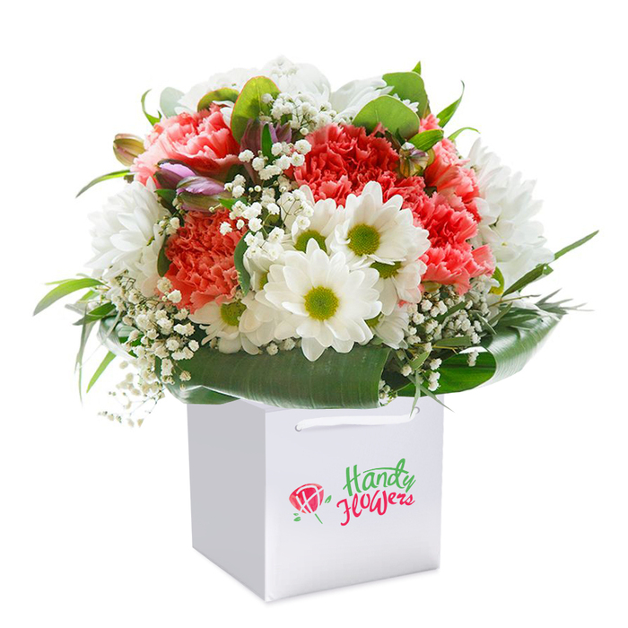 Capturing the artistry of Grove Park's esteemed florists, this vibrant bouquet is thoughtfully arranged inside a modern square white gift bag, ready for delivery across the neighbourhood. Plush coral and peach carnations, their petals delicately ruffled, form the heart of the display, complemented by pure white daisies with striking lime-yellow centers-each bringing a spark of cheerful brightness to the mix. Scattered throughout are soft tufts of baby's breath, lending the arrangement a dreamy, cloudlike softness reminiscent of a stroll through nearby riverside parks. Subtle purple-hued buds, partially closed on the left, add both intrigue and depth, much like hidden floral gems found in Grove Park's community gardens. The bouquet is cradled by an abundance of glossy, deep green leaves that sweep around the base, creating an elegant, full-bodied contour and echoing the leafy tranquillity of Burlington Lane or Fauconberg Road. The rounded profile, careful layering, and interplay of vivid and gentle tones evoke a sense of freshness and thoughtful curation. The clean white bag with secure ribbon handles makes this arrangement suitable for gifting-from celebrating a loved one's birthday to marking a special moment at home. Set against a flawless white background, this detailed image invites the viewer to appreciate the quality, freshness, and neighbourly charm unique to Grove Park Florist's creations.