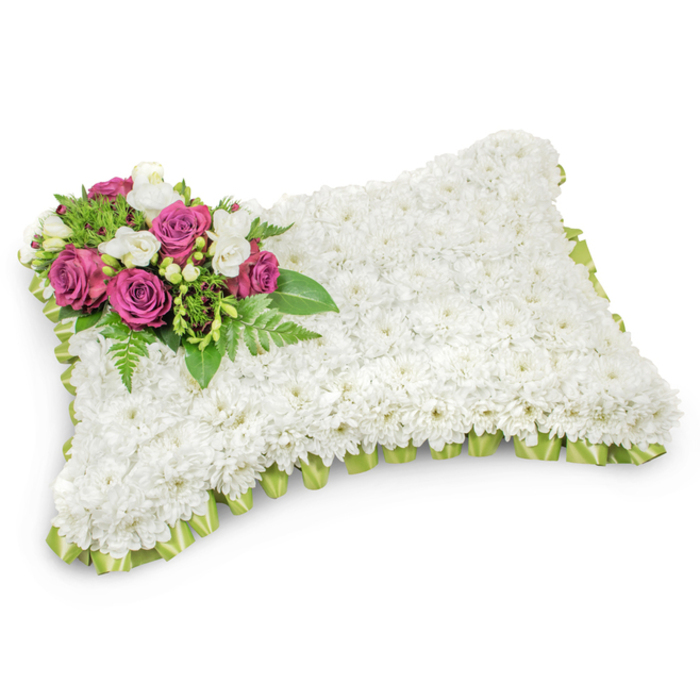 This pillow-shaped memorial arrangement is a study in quiet elegance: hundreds of small white chrysanthemums are placed so closely that they form a continuous, soft-surfaced field across the entire rectangular form, their petals creating a reassuring tactile effect that suggests peace and eternal rest. In the upper-left corner a concentrated spray of rich magenta and deep fuchsia roses becomes the arrangement's emotional centre, the roses' velvety blooms unfolding with dignified grace. Nestled among these are clusters of delicate white freesias or similar buds, which introduce a sweet, imagined aroma and a light visual counterpoint to the intense rose colour. The focal cluster is cushioned by abundant green foliage - glossy leaves and slender fern fronds - that give the tribute structure and a natural, composed look. The edges are finished with a pleated or looped trim of light green satin ribbon that subtly peeks from beneath the chrysanthemum surface, offering a refined border and a hint of fresh colour. Photographed against a plain white background, the pillow's three-dimensional quality is accentuated by a soft shadow, keeping attention entirely on the meticulous workmanship and the soothing palette of white, green, and magenta. As crafted by Grove Park Florist for services in the Grove Park area - whether near the station or in local chapels and green spaces - this traditional tribute conveys respect, consolation, and a gentle honouring of memory, suitable for funerals and graveside remembrances.