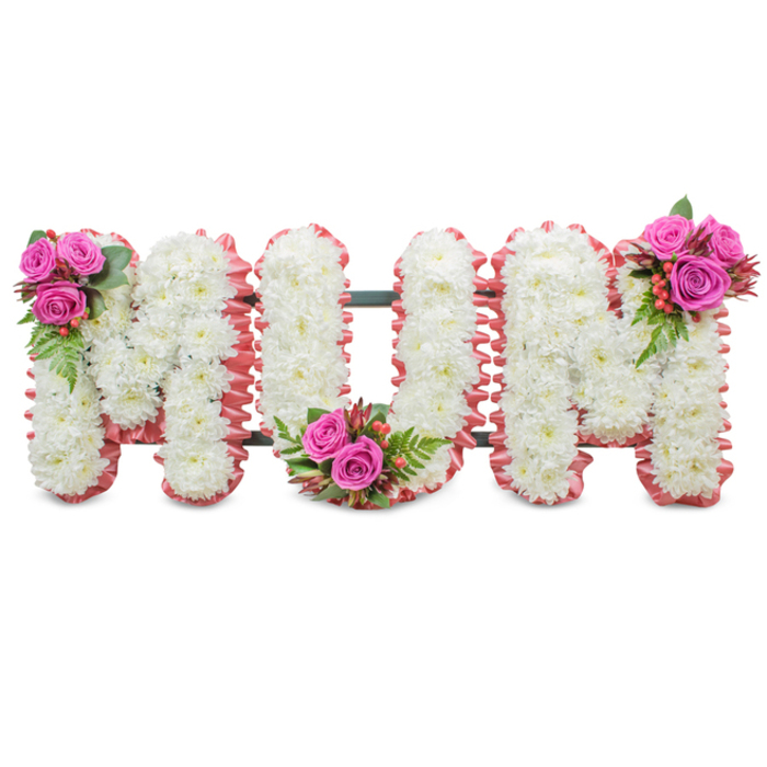 A lovingly composed mourning tribute reads MUM in three large connected letters, each letter densely populated with small white chrysanthemums that produce a refined, almost velvety surface against a stark white background. The composition is edged in a ruffled, pale-pink ribbon trim that softens the bold lettering with an affectionate, almost maternal touch. Between letters a subtle dark green structural frame peeks through, evidence of the careful florist construction that allows this piece to stand steadily at a chapel or cemetery. Three punctuating floral clusters lift the arrangement away from pure white austerity: deep fuchsia-pink roses gathered with ferny green foliage and tiny coral or red hypericum berries appear at the upper left of the first M, at the base curve of the U, and at the upper right of the last M. The juxtaposition of the immaculate chrysanthemums, the plush rose heads and the glossy berries creates a layered tactile quality - crisp, soft and slightly glossy - and suggests a gentle scent of warm roses against a clean, green undertone. This tribute conveys deep respect and affectionate remembrance, intended for memorial services, wakes or graveside placements. For families in Grove Park and surrounding Lewisham neighbourhoods who prefer a classic yet tender floral expression for a mother, this design reads as dignified, intimate and thoughtfully balanced.