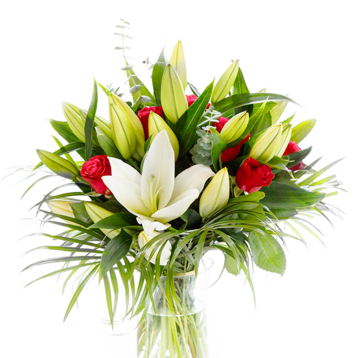This refined bouquet is a study in contrasts: at its heart a large, creamy white Oriental lily blooms fully, its smooth petals curling back to reveal pale yellow stamens and a subtle green pistil, emblematic of purity and calm. Surrounding the central lily, clusters of lime-tinted and buttery-yellow lily buds suggest forthcoming blossoms, adding a hopeful, budding quality to the composition. Interwoven among these are small red rosebuds-deep, velvety, and just beginning to unfurl-whose passionate crimson notes punctuate the arrangement with classic romance. The flowers are set within an abundant framework of greens: dark, glossy leaves that form a structured backdrop; long, slender palm fronds in a lighter emerald that arch and introduce motion; and soft, silvery eucalyptus sprigs that lend a muted, textured contrast and a faint aromatic chill. The clear, cylindrical glass vase reveals the submerged pale-green stems and clean water, anchoring the display with simple elegance beneath bright studio lighting. The white background highlights textures and colour relationships-the lily's satin surface, the roses' plush depth, the eucalyptus's matte sheen-so the bouquet reads as both celebratory and contemplative. Imagine the lilies' powdery, sweet perfume balanced by the roses' richer scent; together they create a bouquet suited to significant moments from anniversaries to sympathetic gestures. This composition would sit beautifully as a centrepiece at a small Grove Park gathering or as a considered gift for someone who loves both classic romance and natural, garden-like texture.