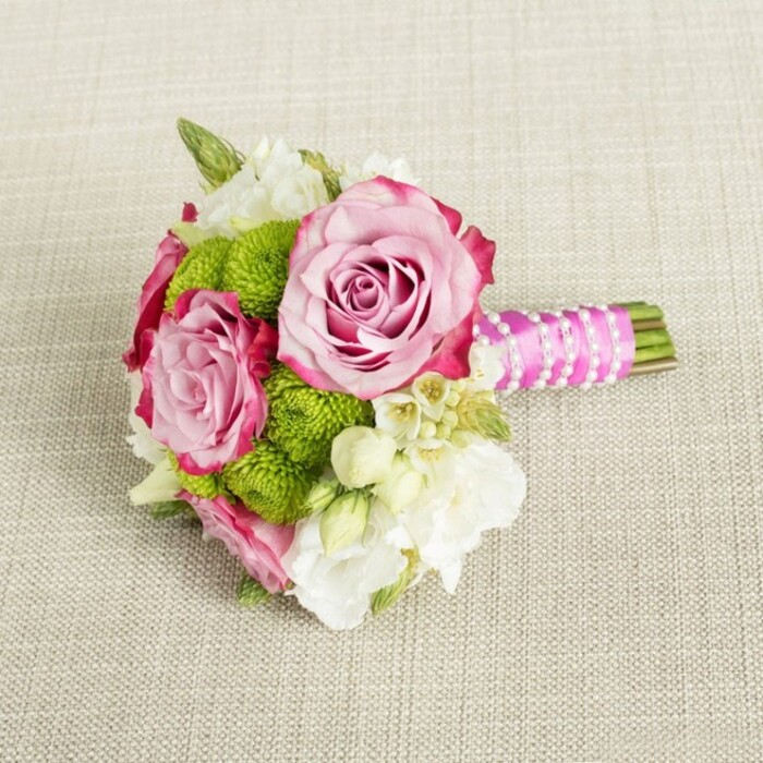 On a softly woven, light tan fabric that could easily be a linen runner on a Grove Park dining table, a compact bouquet rests horizontally, composed with the care of someone preparing for a meaningful occasion. Two prominent roses, fully open yet still holding their structure, draw the eye first. Their petals move from a gentle, almost shell-pink at the centre to deeper fuchsia and magenta tones at the edges, creating a luxurious ombre that makes each bloom look carefully painted. Around and between them, clusters of vivid lime-green button chrysanthemums create cushion-like spheres, their countless tiny petals packed tightly together to deliver both colour contrast and tactile interest. They sit like bright punctuation marks against the softer pinks and creams. Interspersed are creamy white filler flowers - perhaps small lisianthus, freesias, or waxflower - their petal edges soft and slightly ruffled, their petite yellow-tinted centres lending a touch of warmth. A few discreet sprigs of darker green foliage appear at the edges, framing the blossoms without overwhelming them, and hinting at the freshness of stems just cut and tied. The bouquet is bound by hand, its stems trimmed evenly and wrapped in a vivid fuchsia satin ribbon that gleams subtly under the gentle, diffused light. Spiralling delicately over the ribbon, a strand of tiny white faux pearls adds a bridal note, like a necklace adorning a gown. The neutral, textured background keeps all attention on the interplay of pink, green, and white, letting the careful proportions and layering show. This bouquet feels ideal for a bridesmaid walking through Grove Park's leafy streets to a nearby venue, or as a chic centrepiece for a small gathering, reflecting the understated elegance often requested by local clients.