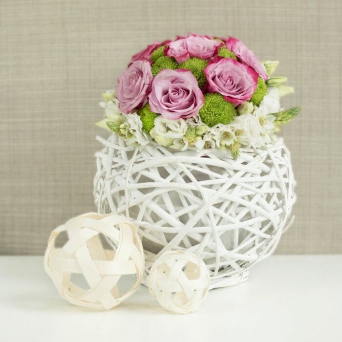 The photograph presents a romantic wedding table arrangement, as though prepared by an experienced local florist for an intimate gathering in Grove Park, not far from the quiet streets around Fauconberg Road. Dominating the centre is a white, round vase shaped from intertwined rattan-like branches, woven into a spherical, open framework that feels both contemporary and organic, its lattice of curved lines catching the soft studio light. From this distinctive vessel rises a perfectly domed bouquet of roses, their colours flowing from gentle blush pink to cool lavender, with some petals kissed by deeper pink edges that add depth and nuance. The roses, mostly in full bloom, swell with layered, velvety petals that create a plush, romantic texture, suggesting both luxury and tenderness. Interspersed among them are bright green button chrysanthemums, their tidy, button-sized heads and fine, densely packed petals bringing texture and a vibrant touch that refreshes the entire palette. Around the lower portion of the dome, delicate white filler flowers weave in and out, forming a feathery fringe that softens the line of the vase and adds a sense of movement, as though the flowers have just been arranged moments before. The arrangement rests on a flawless white tabletop, the smooth surface reflecting soft hints of the vase's white and the blooms' colours. To the left sit two decorative wicker spheres, one larger, one smaller, crafted from light-toned strands that crisscross in open, looping patterns, echoing the vase's style and enhancing the handcrafted, natural theme. Behind everything, a blurred beige fabric backdrop with subtle texture sets a calm, warm tone, ensuring the pink, green, and white hues feel fresh and luminous. The scene captures the quiet elegance of a Grove Park table dressed for a wedding, anniversary dinner, or special brunch, where every detail has been thoughtfully considered.