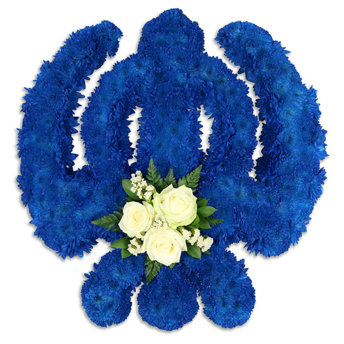 An intricate Sikh Khanda floral tribute is displayed against a clean white surface, its strong lines and curves made entirely from flowers. The emblem's circular chakkar, central vertical blade, and two flanking kirpans have been outlined and filled with countless royal blue chrysanthemums, so closely placed that they merge into a single, velvety-looking field of colour. The petals overlap in gentle layers, catching the light in soft highlights and deeper shadows that accentuate the dimensional shape of the symbol. The rich blue echoes the serene mood of quiet corners around Grove Park, and the overall effect is one of solemn beauty and spiritual strength. At the lower part of the central sword, breaking the expanse of blue, is a carefully positioned accent bouquet. Three pristine white roses rest at the centre, their unfurling petals forming delicate spirals. They are encircled by clusters of tiny white filler blooms, probably baby's breath, whose airy structure adds a touch of lightness. Around this, short stems of dark green foliage create a natural frame, grounding the white against the blue and adding depth to the design. The small posy appears slightly elevated, giving the sense that it has been gently placed upon the Khanda as an offering. The tribute feels fresh and thoughtfully composed, like a bespoke piece ordered for a Sikh celebration of life or remembrance service in Grove Park, where families gather to honour their loved one's faith and heritage. The clean white background keeps every detail in focus, allowing the viewer to appreciate the precise workmanship and the emotional resonance carried by the colours and forms.