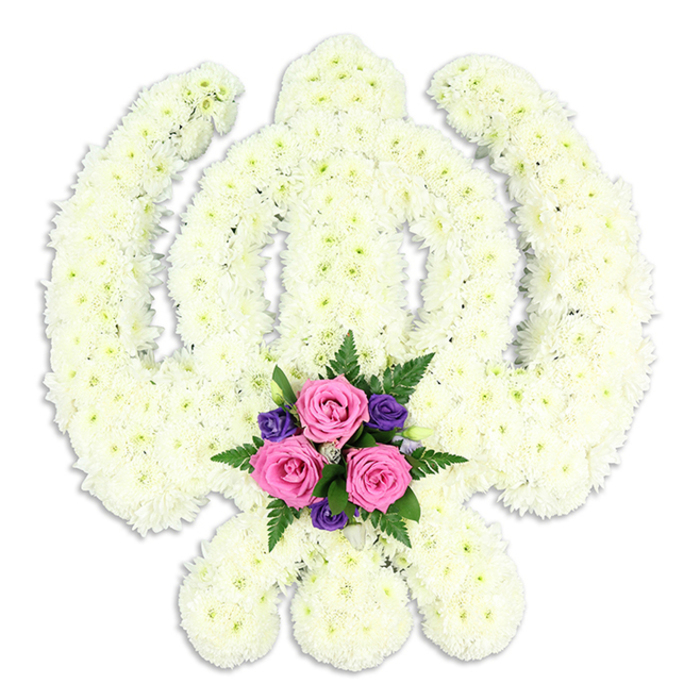A striking Sikh Khanda floral arrangement stands in sharp relief against a bright white background, every line of the sacred symbol translated into flowers. The central Khanda sword rises vertically, surrounded by the round Chakra and flanked by the two curved Kirpans that cross elegantly at the base. The entire surface of this emblem is covered in small, tightly packed white chrysanthemum blooms. Their petals are finely layered and feathery, overlapping so closely that the eye reads a continuous, plush texture rather than individual flowers. The white is soft and creamy rather than harsh, and the even lighting allows gentle shadows to define the intricate shapes, giving the structure a sculpted, three-dimensional look, almost as if the Khanda has been carved from petals.
At the junction where the Kirpans meet, a small accent bouquet brings a thoughtful splash of colour. Three pink roses, their heads full and perfectly formed, sit at the heart of this cluster, their pastel tones warming the cooler whites above. They are framed by several compact, deep purple blossoms, perhaps lisianthus or similar, whose velvety petals deepen the mood without overwhelming the design. Fine green fern fronds and foliage arc outwards around the blooms, introducing freshness and a sense of gentle movement.
This bespoke floral Khanda would feel at home at a Sikh funeral, memorial, or commemorative service in Grove Park and surrounding areas such as Kew or Gunnersbury. One can imagine it gracing the front of a quiet chapel, resting beside a loved one's photograph, or travelling from a family home near Grove Park station, offering a composed, beautiful expression of respect, faith, and enduring remembrance for the person being honoured.