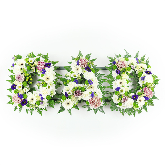 A detailed floral letter tribute spells out the word DAD in three prominent, block-style letters, crafted with the precision and care of a skilled Grove Park florist. The arrangement is displayed against a plain, bright white background, which allows the structure and colour of each letter to be clearly seen and appreciated. The body of every letter is composed of tightly packed white flowers, mostly chrysanthemums and gerbera-like daisies, whose overlapping petals create a soft, cloud-like surface that conveys purity, sympathy, and peace. Interspersed throughout these are smaller white blooms-lisianthus and spray roses-adding variation in size and form, from tight buds to partially open rosettes. This mix of textures gives the arrangement a living, breathing quality, as though each flower represents a cherished memory. The shapes of the D, A, and D are clearly defined by lush green foliage edging and underscoring the flowers. Delicate fern fronds and compact leaves follow the contours of the letters, framing the white blooms with a ribbon of fresh green that provides structure and contrast. To break the monochrome palette and add emotional nuance, the florist has woven in accents of colour: small clusters of bright green button chrysanthemums, deep violet and purple blossoms that might be pansies or spray chrysanthemums, and subtle lavender-mauve roses or carnations. These cooler, elegant tones hint at dignity, eternal love, and quiet reflection. The tribute appears calm and balanced, the kind of personalised DAD lettering often chosen by families in Grove Park for funeral services, memorial gatherings, or graveside tributes along nearby churchyards. It is a floral piece that speaks gently but clearly, honouring a father's life with beauty and thoughtful craftsmanship.