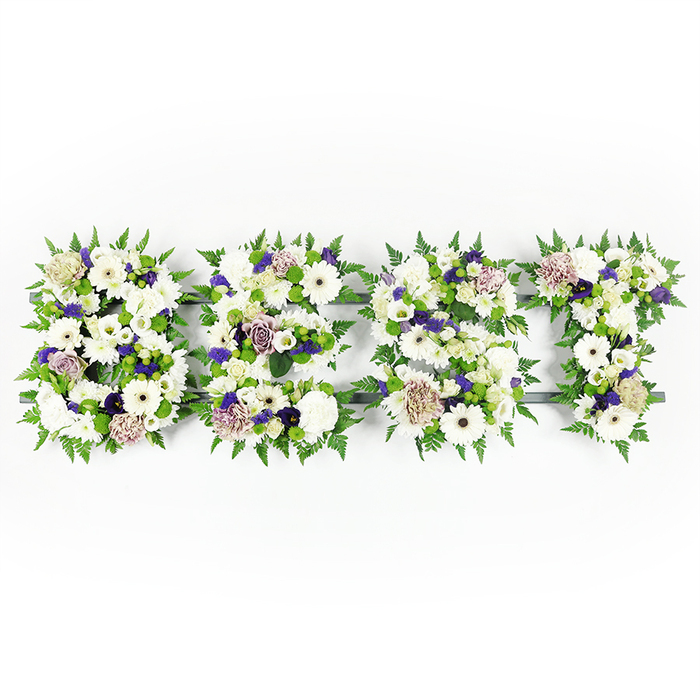 An elegant, horizontal floral display spells BEST in four clearly formed letters, photographed on a clean white background that highlights every shade and texture. Each letter - B, E, S and T - is densely composed of pure white gerbera daisies with their bold, dark centers, layered white lisianthus and full, soft white carnations to create a palette of serene whites; these are subtly warmed by touches of mauve and lavender roses and ruffled carnations, while tiny clusters of deeper violet filler provide quiet depth. The letters are encircled and softened by abundant fern fronds, their feathery green edges lending a natural frame and a sense of movement, and small green button chrysanthemums sit tucked among the blooms to catch the eye with fresh colour. Where the composition is most compact, a dark grey structural base is just visible, a simple armature that defines the letters without intruding on the floral narrative. The lighting is clear and even, giving the flowers a crisp, comforting presence and suggesting the delicate scent of roses and fresh greens. As a floral tribute commonly chosen for memorial services, this piece balances the clarity of the word BEST with layered textures that reward close attention: from the satiny rose petals to the papery gerbera faces and the soft fronds between them. I picture it being chosen by families across Grove Park for wakes and services at nearby community halls or churches, a personal and dignified expression of love, respect and remembrance crafted with steady hands and local care.