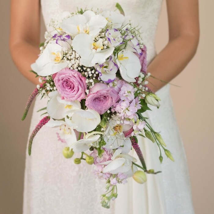This photograph highlights the tender artistry of a Grove Park wedding, focusing on the bride mid-frame as she holds a showstopping cascading bouquet. Her white or ivory gown, featuring gently shimmering lace, offers an exquisite backdrop that sets the stage for the florals' soft hues and abundant textures. The bouquet itself unfurls in a magnificent waterfall: central white Phalaenopsis orchids, their velvety petals and ephemeral gold centers glistening in the light, preside over roses blushing in lavender and pink. Some roses bloom with rich fullness, others peek shyly from beneath airy sprays of gypsophila-whose tiny blossoms float like clouds across the floral landscape. Slender, bright magenta amaranthus trails and curled green foliage provide contrast, movement, and a modern touch, evoking the lush greenery found along Strand-on-the-Green paths. Smaller lavender and sweet pea blossoms add subtle texture and depth, and unopened buds invoke hope and renewal. The colors-white, pink, purple-blend in harmonious sophistication, threading together the timeless with the contemporary. The bride's careful cradle and the subtle, muted backdrop focus attention squarely on the bouquet, a symbol not only of personal joy but of neighborhood pride. Perfect for those celebrating at community parish halls or riverside pavilions, this arrangement encapsulates the refined style and heartfelt service at Grove Park Florist.