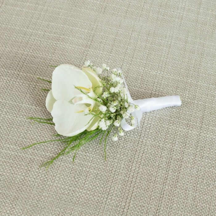 A meticulously crafted white orchid boutonniere is photographed resting on a light beige, woven fabric that looks like natural linen, the kind of understated textile you might see in a stylish Grove Park home. The neutral backdrop, with its gentle texture and warm, sandy tone, allows every detail of the floral piece to stand out. At the centre of the arrangement sits a luminous Phalaenopsis orchid bloom, its petals wide, smooth, and almost porcelain-like in their purity. The petals unfurl in a graceful arc, leading the eye toward the orchid's throat, where a hint of pale yellow glows softly, giving the flower a sense of freshness and living warmth. Around this central orchid, a generous cluster of baby's breath adds a halo of tiny white blossoms. Each gypsophila floret is no larger than a pinhead, yet collectively they create a cloud-like ring that softens the edges of the boutonniere, suggesting romance and lightness well suited to a wedding at a nearby Grove Park venue or a chic registry office in West London. Nestled beneath and between these blooms are fine, feathery strands of bright green foliage, reminiscent of delicate fern fronds, that bring a gentle movement and a touch of garden woodland character, as if picked from the greenery near Chiswick House and Gardens. The stems are drawn tightly together and bound in a strip of white satin ribbon, matte and smooth, wrapped with practised care so no loose ends show. A small, tidy bow sits where the stems meet the blooms, indicating the point at which the boutonniere would be pinned to a lapel. The lighting is calm and balanced, with no harsh glare, so the white tones remain soft rather than stark. Overall, the image evokes quiet elegance and timeless sophistication, the sort of boutonniere a groom or groomsman might order from Grove Park Florist for a classic, refined celebration in the local area.