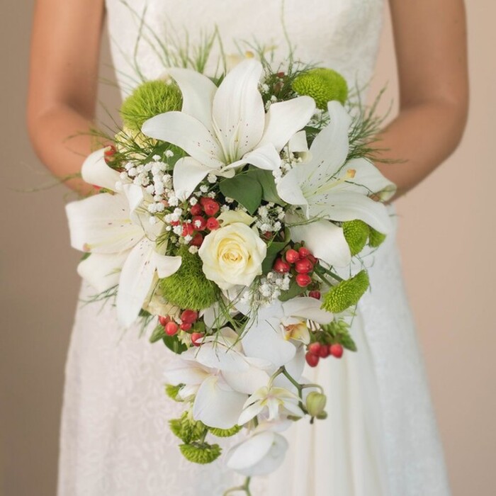 The image frames a bride from the waist up in Grove Park, gently holding a lavish cascading bouquet that embodies elegance, vibrancy, and local charm. Lofty white lilies command attention at the bouquet's centre, their trumpet forms adorned with delicate reddish-brown speckles, creating a sense of refined grandeur. At the heart lies a classic creamy white rose, its spiralled petals closed in a poetic swirl symbolizing enduring love. Featuring prominently among the white blooms are vivid lime-green cushion mums, offering a dynamic splash of colour inspired by the lush foliage of Chiswick's and Grove Park's landscaped gardens. Baby's breath, with its frothy clusters, imparts a soft, cloudlike border, effortlessly complimenting the bouquet's more substantial blooms. Bright red hypericum berries tucked between the flowers add a lively, festive touch-an homage to the spirited celebrations that unite the local community. Trailing elegantly downward are the delicate, snowy white blooms of Phalaenopsis orchids, whose presence defines the bouquet's signature waterfall shape and brings motion to the composition. Fine strands of grass-like greenery and subtle foliage cradle the ensemble in a naturalistic embrace, reminiscent of an outdoor gathering along the Thames Path. The bride's hands and the gentle lace of her ivory dress serve as a romantic canvas, softly blurred to keep the spotlight on the bouquet's artistry. With each fresh flower selected for its beauty and resilience, this design is the perfect choice for Grove Park couples seeking a memorable, bespoke statement on their special day.