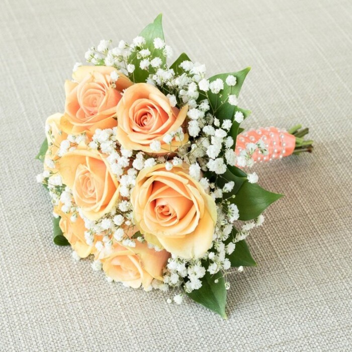 A true expression of Grove Park's refined floral traditions, this bouquet is elegantly positioned atop a lightly textured linen, the kind one might find in welcoming local venues near Baring Road. Seven lush roses are the bouquet's focal point, each in a radiant gradient from warm peach to pale apricot, their tightly wound centers gracefully unfurling outward to suggest life and promise. Among the roses, baby's breath blooms in white, airy clusters-ethereal as soft spring clouds rolling above the South East London skyline. Sprigs of deep green foliage peek through, lending organic depth and harmony. Gathered into a balanced, classic round shape, the bouquet is bound by a smooth, peachy satin ribbon, its delicate tone perfectly matched to the roses. Spirals of tiny faux pearls embellish the ribbon-wrapped handle, adding a touch of vintage celebration. At the base, neatly trimmed stems are visible beneath the wrap-a mark of freshness and professional floristry. Diffuse, gentle lighting showcases the subtle variation of petals and the gleaming pearls, highlighting every comforting nuance. Ideal for Grove Park weddings, anniversaries, or as a thoughtful gift delivered across Bromley and beyond, this arrangement weaves together the grace of handcrafted design, the richness of local greenery, and the promise of new beginnings, all against the quiet backdrop of the community's cherished traditions.