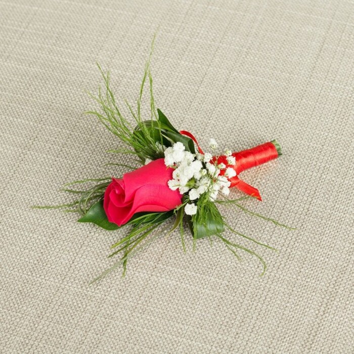 A single-stem boutonnière is shown laid at a gentle angle across a light, oatmeal-beige fabric backdrop, whose fine weave and soft creases suggest a natural linen or cotton often used in calm, contemporary interiors around Grove Park. At its centre, a tightly closed rosebud in a rich fuchsia or deep rose-pink tone draws the eye, its velvety petals coiled inward so that the bloom looks full of contained energy, poised between bud and blossom. Around this central flower, clusters of delicate white baby's breath nestle close, each tiny flower a crisp, star-shaped accent that sets off the saturated colour of the rose and introduces a whisper-light texture to the composition. From behind the rose, slim, grass-like sprays of bright green foliage emerge and fan outward, creating a soft halo that gives the arrangement height and a sense of natural movement, as though plucked from a garden walk near Grove Park station and arranged with practiced care. Deeper green leaves, slightly broader and more solid, sit at the base, adding structure and visual weight so the design feels balanced rather than fragile. The stems are carefully bound in a smooth red satin ribbon, wrapped several times to create a neat, cylindrical handle that ends in a tiny loop or subtle fold, a hallmark of attentive craftsmanship you might expect from a local specialist like Grove Park Florist. Gentle, even lighting picks up the sheen on the ribbon, the faint texture of the background fabric, and the fresh, almost dewy quality of the petals and foliage. Overall, the image conveys quiet romance and thoughtful detail, suggesting a groom's lapel flower or formal buttonhole prepared for a wedding or prom celebration in and around Grove Park, ready to be pinned just before the photographs begin.
