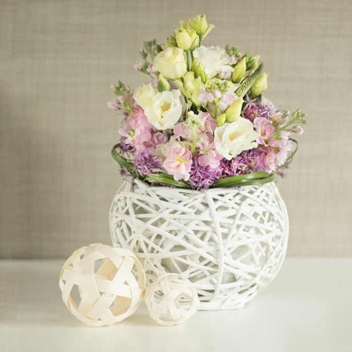 This lush floral tableau, arranged by a skilled Grove Park florist, perfectly captures the tranquillity and refined charm that the area is known for. Centrally displayed in a white, spherical vase that seems almost to float on a sunlit table, the arrangement bursts with pastel-hued eustoma-ivory, pale yellow, and soft pink-each flower's ruffled edge gracefully catching the day's light. Spires of delicate pink stock (matthiola) lift the eye skyward, while at the base, a tapestry of rich purple fillers-possibly allium or aster-intertwines with verdant foliage, bringing both depth and a gentle pop of colour. The vase itself is a modern showpiece, fashioned from open-work rattan or dried painted willow branches, its airy construction allowing light and shadow to dance across the flowers and table below. To the left, two decorative spheres, carefully woven from pale strips of rattan, echo the vase's intricate artistry, uniting the entire arrangement with the organic, welcoming spirit of Grove Park homes. The background offers a soft, neutral backdrop, suggesting the gentle palettes found in the area's period interiors. Bathed in soft daylight, the image evokes feelings of freshness and quiet celebration, making it ideal for Grove Park wedding receptions, sophisticated living room centrepieces, or as an inspiration for spring home décor with a local touch.