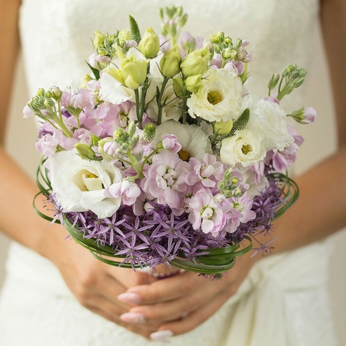 Captured in luminous detail, this close-up celebrates the union of craftsmanship and locality found in Grove Park bouquets. At its core, white Lisianthus blooms fan open, their ruffled petals and soft yellow-green centers glowing with purity and hope. Amidst them, frothy pastel stock flowers in pinks and lavenders interlace, evoking the gentle palette of wildflowers growing beside Grove Park's tree-lined walks. The design is further enriched by slender, unopened buds and loops of glossy bear grass, weaving subtle structure and freshness through the arrangement, much like the meandering pathways of Grove Park's green avenues. Anchoring the entire composition, a distinctive halo of deep purple, starry Allium flowers wraps the base, offering a geometric, modern foil to the bouquet's soft lines. The bride's hands, perfectly manicured in pale tones, lovingly support the partially wrapped stems, conveying both anticipation and affection. In the softly blurred background, delicate hints of a traditional lace wedding dress elevate the sense of occasion without distracting from the bouquet's pastel artistry. Perfect for marking a wedding in one of Grove Park's historic venues or intimate gatherings, this bouquet speaks of elegance, local beauty, and the joy of new beginnings.