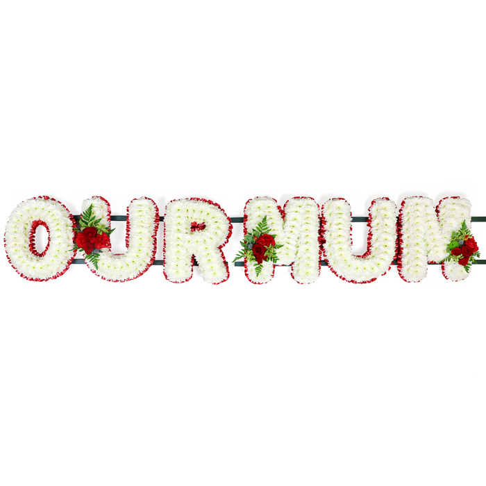 Set against a bright, uncluttered white background, this floral memorial arrangement spells out OUR MUM in large, three-dimensional block letters that appear both strong and tender. Each individual letter is generously covered in countless small, pristine white chrysanthemum-like florets, pressed closely together to create a thick, cushiony layer of petals. The texture feels soft and cool, as if the flowers have just been arranged by a careful florist in Grove Park preparing for a same-day funeral service. The uniform white surface suggests purity and serenity, similar to the peaceful atmosphere of a quiet corner of Grove Park or a tranquil spot in nearby Dukes Meadows. Around the perimeter of each letter runs a precise border of deep red flowers, possibly carnations or compact spray blooms, which traces the shape of every curve and corner with clarity. This red edging makes the lettering stand out clearly and adds a traditional touch of dignity. Behind the blossoms, a dark green structural bar links the letters, creating a single, cohesive tribute ready to be placed at a church, crematorium, or graveside along Burlington Lane or by the Thames. Interspersed among the white petals are three standout clusters of deep red roses accompanied by glossy green foliage and airy ferns. One cluster rests between the O and U, another nestles between the first M and the U, and a final group accents the right side of the last M. These roses introduce depth and emotional warmth; their rich red colour speaks of unwavering love, remembrance, and gratitude for a cherished mum. The whole OUR MUM design feels gentle yet substantial, a classic funeral letter tribute reflecting the quiet strength of family bonds in Grove Park.
