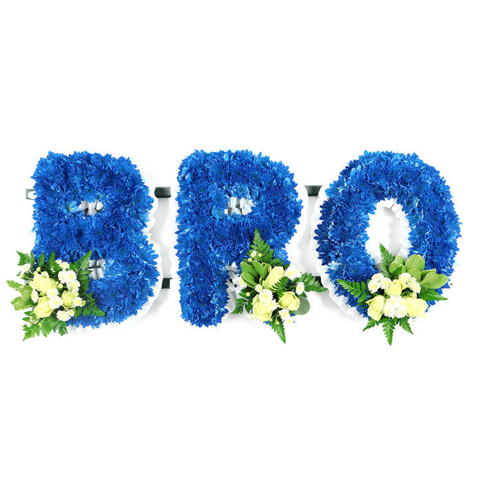 In a crisp, neutral studio setting on a stark white background, a three-part funeral tribute spells the word BRO in large capital letters, each letter lovingly formed from a dense carpet of royal blue chrysanthemums. The clustered blue florets create a sumptuous, velvety texture and precise contours; the white supporting frame or floral foam is subtly visible around internal cut-outs and edges, confirming the letters are freestanding. Each character is softened with an elegant accent: small sprays of pale ivory and creamy-yellow spray roses, some tight and others gently opening, are grouped with petite white filler blooms and artfully arranged foliage - glossy salal-like leaves and delicate, airy fern fronds - placed at the lower-left of the B and the lower-right of the R and O. The interplay of deep blue, warm cream and fresh greens is harmonious, while the neutral lighting lets the colours breathe and reveals the tactile density of the chrysanthemum heads. This is a considered memorial design, quiet in tone yet unmistakably personal; the blue expresses calm and loyalty, the cream roses evoke tenderness and consolation. As an experienced local florist, Grove Park Florist produces memorial pieces like this for families around Grove Park, Elmstead Woods and Downham, often for services at nearby churches, community halls or small chapels - its clean silhouette and gentle scent make it fitting for placement beside a photo board, on a stand in a chapel, or at a graveside. The arrangement reads as both a formal tribute and a private message of enduring brotherhood.