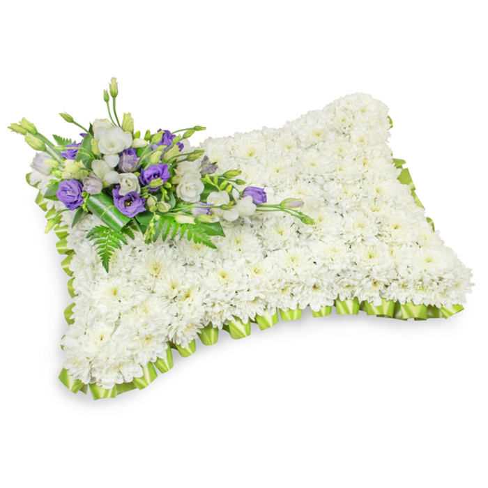An exquisitely made floral sympathy pillow, presented like a small, serene bed of blooms, is formed entirely from hundreds of tiny white chrysanthemum heads that have been densely positioned to create a uniform, plush surface. The arrangement holds a soft, pillow-like volume with rounded corners and a slightly raised profile; under gentle studio lighting the piece casts a subtle shadow that highlights its three-dimensionality and meticulous construction. A fine, light lime-green ribbon trim is pleated around the full perimeter in delicate, overlapping loops, the cheerful yet subtle green providing a crisp frame to the tranquil white mass.

Across the upper-left corner rests an elegant cluster of accent flowers and greenery: ruffled white lisianthus sit beside deeper purple lisianthus, some blooms fully open and others still furled as buds, offering a poignant contrast in tone and texture. Feathered fern fronds and broad dark leaves are woven through the cluster, and a deeper green leaf wrap at the base secures the stems neatly while lending an organic finish. The bouquet is scented faintly - a gentle clean aroma of chrysanthemum with the soft, floral hush of lisianthus - appropriate for quiet remembrance.

Prepared by Grove Park Florist for families across Grove Park and neighbouring Downham and Lee, this cushion-style memorial is intended as a respectful tribute for funerals, memorial services, or graveside ceremonies. It reads as a peaceful, comforting emblem of condolence, designed to offer gentle solace when gathered to remember a loved one.