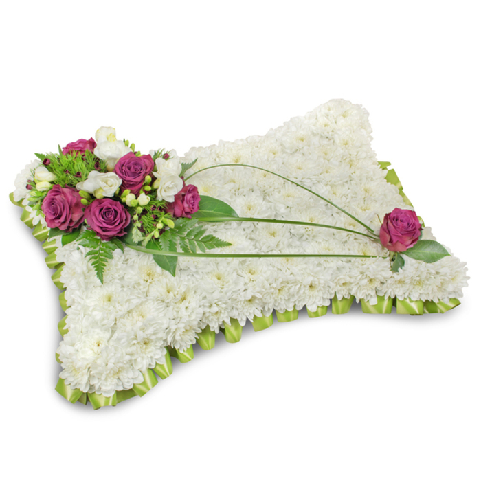 A traditional pillow-shaped funeral tribute photographed against white to emphasise its solemn beauty and serenity for families in Grove Park and the surrounding neighbourhood. The cushion is completely covered in small, immaculate white chrysanthemum blooms, densely formed so the surface becomes a plush, cloudlike expanse that suggests rest and purity. The lower edge is finished with a continuous pleated band of pale lime-green satin ribbon, folded to create a scalloped, ruffled border that frames the white surface with a whisper of colour. Anchoring the design in the upper left is a compact cluster of deep magenta roses - lush, velvety blooms - interspersed with smaller white freesia or delicate lisianthus, which introduce a soft, fragrant lift and a sense of fragile grace. Fresh green foliage, including delicate fern fronds and glossy leaves, fans out beneath the cluster, adding structure and a natural backdrop. Across the pillow's face three slender, arching blades of bear grass traverse the chrysanthemums, their elegant lines drawing the eye toward the lower right where a single magenta rose echoes the upper grouping and completes the composition. The interplay of textures - the chrysanthemum's tight petals, the roses' velvet, the satin ribbon's sheen, the fern's featheriness - is captured in gentle light that deepens the tribute's contemplative mood. Ideal for services at local churches or for delivery near Grove Park station and the nature reserve, this arrangement communicates sympathy, comfort, and a respectful farewell.