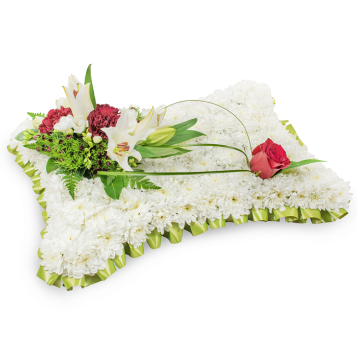 A classic funeral cushion rendered in flowers: a neat rectangular pad entirely covered by hundreds of small white chrysanthemums whose closely packed petals form a soft, plush plane reminiscent of a fabric pillow. The chrysanthemums' uniform white creates a feeling of purity and calm, while a pale lime-green satin ribbon, pleated into a subtle frill, neatly encircles the edge and gives the piece a finely finished, respectful border. The photograph's clean white background highlights the arrangement's textures and gentle shadows, allowing the craftsmanship to read clearly.

Anchoring the design on the upper left is a composed cluster of white Oriental lilies - two in full bloom, their pale petals opening to reveal warm yellow stamens, with a closed bud nearby - together with richly coloured burgundy spray carnations that bring warmth and contrast. Varied green foliage, including feathery ferns and broader leaves, provides a lush backdrop and natural depth. From that cluster a thin, arching strand of salal leaf or decorative wire extends across the chrysanthemum surface and culminates on the upper right with a single deep red rosebud, its velvet petals just beginning to unfurl. The lilies introduce a soft fragrance, while the rosebud and darker carnations add a note of enduring love.

This arrangement reads as a timeless expression of sympathy suitable for services in and around Grove Park - an elegant, comfort-giving tribute often chosen for wakes or memorials at local churches and chapels near Chinbrook Meadow and the surrounding streets.