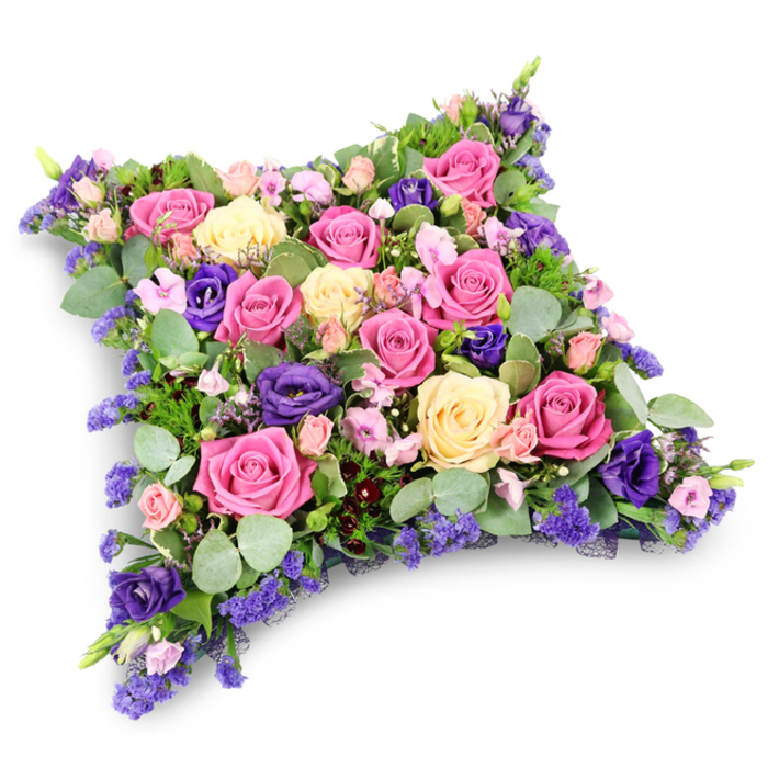 A richly detailed floral cushion, intended as a sympathy or funeral tribute, is shown in full view against a pure white background with only a faint shadow beneath to hint at its weight. The cushion takes on a soft, rectangular shape with smoothly rounded corners, recalling the comforting form of a pillow laid lovingly at rest. Its entire surface is densely planted with roses and complementary flowers in a palette of pink, cream, yellow, and purple. Bold, fuchsia-toned roses create vibrant focal points, their open blooms revealing curled petals layered like fine fabric, while gentler blush pink and creamy pale yellow roses lend a more subdued, tender character. Between these, deep purple lisianthus flowers appear in small clusters, their velvety, ruffled petals contributing a refined elegance that feels appropriate for a farewell service at a Grove Park church or crematorium. Fine sprays of tiny lavender and lilac filler blossoms, likely statice or limonium, fill the smallest gaps, adding a delicate haze of colour that unites the arrangement. Supporting the flowers is a bed of mixed greenery: rounded silver-green eucalyptus leaves provide a soft, contemporary touch, their matte surfaces contrasting with the gloss of the rose petals, while darker, more feathery foliage lines the edges, emphasising the gentle curve of the cushion. The lighting is gentle and even, revealing subtle shadows between the blooms and enhancing their three-dimensional form without harshness. The tribute conveys an atmosphere of peace, remembrance, and enduring affection, as though created for families in Grove Park who wish to send a floral message of comfort and respect when words feel too small.