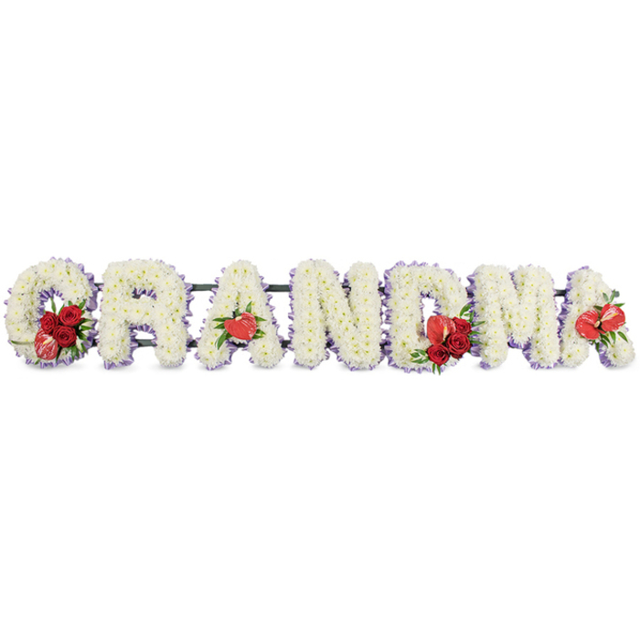 A beautifully detailed GRANDMA floral arrangement stretches horizontally across the image, captured against a clean white background that draws attention to each individual bloom. The six capital letters are sculpted from clusters of white cushion chrysanthemums, or similar small-headed flowers, packed so densely that the surface appears soft and velvety with a gentle, almost luminous sheen. The flowers are arranged evenly and carefully, showcasing the skill of an experienced local florist familiar with heartfelt tributes in Grove Park. Along the perimeter of every letter, a narrow edge of pale lavender or soft purple backing gently frames the white petals, creating a delicate outline that adds depth and a sense of tender calm. Interspersed among the letters are striking red floral accents that inject warmth and emotion: the G holds a small bouquet of deep red roses, their layered petals opening generously, seated beside a distinctive red-and-white variegated calla lily whose curved form leans outward. The first A is embellished with a single red anthurium at its base, its waxy, heart-shaped spathe catching the light. At the centre, the M carries another group of rich red roses together with an additional red anthurium, while the final A mirrors this detail with one more anthurium bloom, visually tying the tribute together. A dark green stand or framework runs discreetly behind the letters, supporting their weight and ensuring a tidy presentation that would sit comfortably on a stand at a Grove Park funeral service or crematorium chapel. The overall effect is one of reverence and gentle affection, a carefully personalised floral message dedicated to the memory of a much-loved grandmother.