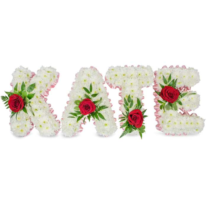 This elegant memorial arrangement presents the name KATE in bold block letters, each letter meticulously formed from a dense mass of crisp white chrysanthemums that create a soft, almost cloud-like texture. The faces of the letters are edged with a pale pink pleated ribbon that follows every contour, lending a gentle blush to the stark white composition and a finished, refined silhouette against the clean white backdrop.

Each letter bears a single deep red rose as its visual centre, each rose nestled within abundant green foliage composed of broad glossy leaves and slender ferny fronds that add layered texture and a sense of life. Placement is carefully considered: the red rose on the K is positioned on the lower part of the left vertical stroke; the A's rose crowns the top of its triangular peak; the T's rose is set at the base of the left side of the horizontal crossbar; and the E's rose sits upon the middle horizontal bar. The subtle scent of rose threads through the arrangement while the chrysanthemums provide a restrained, classic sympathy tone. Designed for use as funeral flowers or a memorial name tribute, this composition conveys heartfelt remembrance - a clear, dignified way to honour someone's life, suitable for display at services near Chinbrook Meadows, local churches, or community venues around Grove Park.