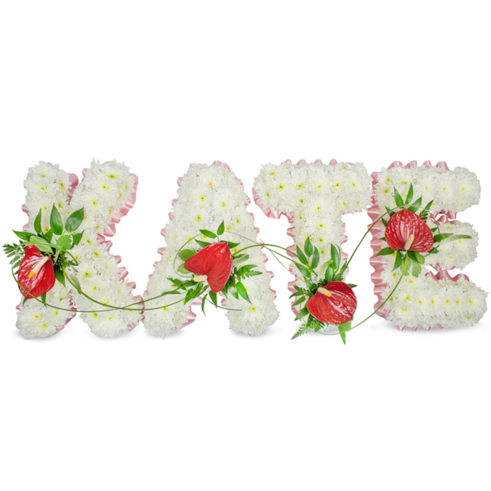 This elegant memorial display spells the name KATE in large, capital floral letters, each formed from a dense, uniform carpet of small white chrysanthemums that create a soft, tactile surface evocative of peace and reverence. The outer edges of the letters are finished with a pleated light pink ribbon, its gentle hue delicate against the pure white blooms and adding an artisan's finishing touch. Nestled into the chrysanthemum base are concentrated groupings of vibrant red anthuriums; their glossy, heart-shaped spathes and prominent spadices stand out as warm, emotional highlights amid the serene palette. Rich green foliage - broad glossy leaves, airy fern fronds and smart sprigs of ruscus - is woven through and around each letter to provide contrast, depth and a natural framework. Slender green tendrils and vines arc between letters, visually sewing the individual forms into a single, flowing tribute. Under soft, directional lighting the composition casts a subtle drop shadow on the clean white background, enhancing its three-dimensional presence and making it easily readable at a funeral or memorial display. The arrangement feels intimate and composed, the craftsmanship reflecting longstanding funeral floristry traditions common to local services in Grove Park, Lewisham and neighbouring Lee. One can almost sense the faint, green fragrance of fresh foliage mingling with the mild, powdery scent of chrysanthemums - an understated aroma suited to quiet reflection. This is a bespoke, comforting tribute intended to honour memory with tenderness and dignified beauty.