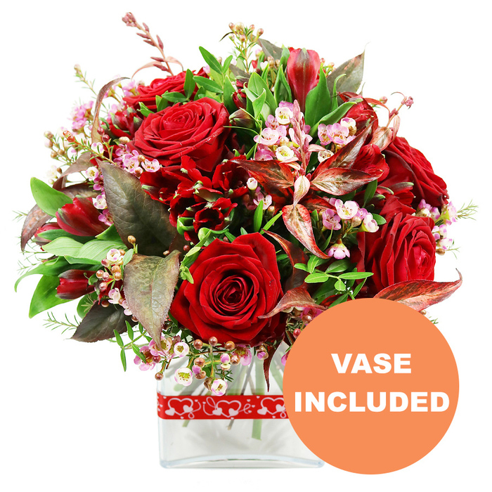 A lavish floral arrangement in rich shades of red and green is displayed against a pristine white background, evoking the sense of a thoughtfully chosen bouquet awaiting delivery to a cosy home in Grove Park. The design is rounded and full, with several velvety red roses acting as the focal point; their petals, some tightly wound and others unfolding, create layers of texture and express a spectrum of emotion from anticipation to fully blossomed love. Threaded through the roses are clusters of smaller deep crimson flowers, akin to alstroemeria or similarly shaped multi-petalled blooms, which fill the gaps and provide a rhythmic repetition of colour and form. Between these more dominant elements, delicate sprays of tiny pale pink and white filler flowers-resembling waxflower or limonium-appear like scattered stardust, their minute blossoms and branching stems lending a soft, airy lightness that offsets the intensity of the reds. The entire arrangement is encircled by an abundance of foliage: fresh green leaves in varying shapes and tones frame the blooms, suggesting vitality and freshness, while pockets of distinctive burgundy and reddish-brown leaves with fine veining add depth, warmth, and a slight touch of drama. The bouquet is set in a clear, modern rectangular glass vase, its transparent walls revealing the clean lines of the stems and the clarity of the water, giving the design a contemporary, refined edge suitable for dining rooms, coffee tables, or sideboards along the quiet residential streets of Grove Park. Wrapped neatly around the vase is a red ribbon patterned with small white hearts, quietly referencing love, commitment, and the special moments this bouquet might celebrate-from anniversaries and Valentine's Day to heartfelt apologies or thank-you gestures. In the bottom right corner of the image, a prominent circular orange label with bold white uppercase text reads VASE INCLUDED, making it immediately clear that this floral gift arrives complete, ready to display straight out of the box.