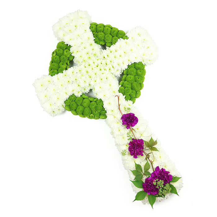 The image shows an elegant Celtic cross floral tribute, carefully arranged and set against a pure white studio backdrop that enhances the clarity of the design. The cross appears slightly angled, giving a sense of flow while remaining strong and composed. Its main structure is built from tightly clustered white chrysanthemums, each small bloom contributing to a soft, cushion-like blanket of petals. These white flowers fill the vertical and horizontal arms as well as parts of the circular centre, creating a calm, serene base. Defining the traditional Celtic ring and interlaced lines is a vivid band of lime-green button chrysanthemums, placed in continuous, carefully shaped rows. Their compact, rounded heads give the arrangement crisp outline and visual rhythm, making the Celtic motif immediately recognisable. The balance of white and green brings to mind the calm of local green spaces near Grove Park, such as the riverside walks where many families find quiet moments of reflection. Flowing down the lower arm of the cross, three distinct clusters of deep purple carnations provide a rich, heartfelt accent. Their densely layered petals are slightly ruffled, introducing a velvety depth that stands out against the smoother chrysanthemums. Slim strands of greenery and a few naturally curling stems wind gently around these purple groups, adding movement and a touch of wild, garden beauty. The lighting is soft and even, allowing the viewer to appreciate the textures and three-dimensional quality of the cross without distraction. This floral tribute feels both traditional and personal, suitable for a funeral, memorial, or celebration of life in Grove Park, particularly when there is a wish to reflect Celtic heritage, Christian faith, or a deep appreciation for nature.