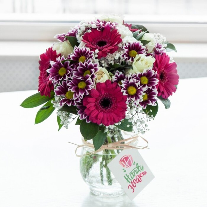 A lush, vibrant bouquet is carefully staged in the centre of the frame, resting in a low, clear glass vase that reveals the freshly cut green stems gathered neatly within. The glass has a gentle texture that catches the light, and the whole arrangement sits against a bright, softly blurred white background that feels like a sunlit window in a quiet Grove Park kitchen. At the heart of the design are striking gerbera daisies in saturated fuchsia and magenta tones, each with a velvety dark centre that pulls the eye inward and gives the bouquet a confident focal point. Around them cluster multi-layered chrysanthemums, their petals tipped in deep purple and fading gracefully to white, forming daisy-like faces with golden yellow centres that add warmth and cheer. Nestled amongst these are creamy white roses, some petals unfolding in full, cup-shaped blooms while others remain in elegant, tight buds, suggesting new blossoms and bringing a soft, romantic dimension to the composition. Delicate sprays of white baby's breath float gently through the spaces between larger flowers, adding fine, lace-like texture and a sense of airiness that keeps the arrangement from feeling heavy. Glossy deep green leaves emerge at the base and along the sides, framing the colourful blooms and giving a natural, garden-fresh feel, as though the flowers had just been gathered from a well-tended border near Grove Park's leafy avenues. The neck of the vase is wrapped with rustic twine tied into a modest bow, from which a small white rectangular tag bearing a simple floral symbol dangles, ready for a personal note. The overall mood is joyful and celebratory, ideal for a birthday breakfast table, a thank-you gift, or same-day delivery from a trusted local florist across the wider Grove Park area.