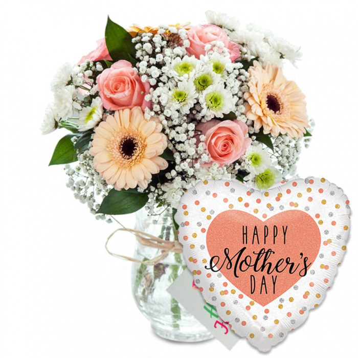In this image, a thoughtfully arranged Mother's Day gift set is displayed against a smooth white background, allowing every curve of flower and foil to stand out. On the left, a clear glass vase anchors the composition. Its gently rounded form is unadorned except for a light band of rustic twine wound around its neck, tied in a modest bow that conveys an artisan, hand-finished touch so familiar to customers of Grove Park Florist. Rising from the vase is a lush bouquet, shaped into a full, rounded cluster. The colour palette focuses on soft, romantic hues: several peach and blush-pink roses open in layered spirals, their petals slightly ruffled at the edges, adding texture and a sense of life. Around them, peach-toned gerbera daisies, each with a dark chocolate centre, fan out their neat, uniform petals, bringing a cheerful, contemporary note to the design. Wispy sprays of white baby's breath thread between these larger blooms, forming a light, cloudlike veil, while compact white chrysanthemums dotted with pale green centres fill the gaps and give the arrangement a sense of completeness. The greenery is deep and glossy, arching around the blooms to frame them, reminiscent of the lush hedges and trees that line Grove Park's quiet residential avenues. Behind the flowers, a corner of a small white card is just visible, with hints of pink and green writing that likely spell out a Mother's Day greeting, as personal and intimate as a note left on a breakfast tray. To the right, slightly overlapping the base of the vase, a large heart-shaped foil balloon leans gracefully. Its surface is bright white, sprinkled with a lively pattern of metallic gold and silver polka dots. In the centre, a peach-coloured heart carries the words HAPPY Mother's DAY in bold black typography, with Mother's rendered in elegant, flowing script. The overall effect is of a polished yet heartfelt gift, ready to be delivered to doorsteps across Grove Park and beyond.