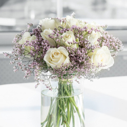 A calm and dignified bouquet of sympathy flowers, featuring creamy white roses and soft purple filler blooms, is shown in a detailed close-up that highlights both structure and emotion. The roses are the stars of the arrangement, their velvety petals unfolding from tight spiral centres into full, open faces that exude purity and tenderness, their colour sitting between pure white and gentle ivory. Intertwined with these generous blooms are fine shoots of tiny lavender and lilac blossoms, probably statice, forming airy tendrils and small clouds that soften the stronger lines of the roses and add a subtle wash of colour. All of the flowers are arranged in a simple, tall, cylindrical glass vase, so clear that each straight, green stem can be seen clearly, reaching down into fresh, transparent water. The vase stands on a bright white surface, which echoes the tone of the roses and catches the light in such a way that faint reflections shimmer beneath the arrangement. Behind it, the background is softly out of focus, a blend of white and pale grey that suggests a quiet, light-filled room in Grove Park, perhaps near the calm of the Thames path or one of the area's tree-lined streets. The overall effect is tranquil and respectful, making this bouquet suitable for expressing heartfelt sympathy or offering a gentle reminder of support. The image feels like a moment of stillness in a busy day, a floral pause for contemplation and remembrance, shaped by the hands of a local Grove Park florist who understands the nuances of saying goodbye.