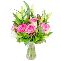 A carefully balanced floral composition of rich pink roses and white lily buds fills a clear glass vase, the whole set against a pristine white background to emphasise colour, form and freshness. Prominent are several fully opened roses, their velvety petals layered in shades from deep fuchsia to softer blush, each bloom exuding the warm, familiar scent of classic garden roses. The white lilies stand like graceful notes between them - many as closed, elegant buds promising to open, a few just unfurling to reveal smooth, fragrant trumpets that contrast with the rounded rose heads. Surrounding these focal blooms is abundant greenery: broad, glossy leaves give weight and structure while finer sprigs introduce lightness and movement; peppered throughout are small, darker reddish-brown accents that add depth and visual interest. The vase is simple and transparent with a subtle tapered silhouette; clear water reveals trimmed stems arranged with care, a detail every florist attends to for longevity and presentation. Photographed in bright, clean light, the bouquet feels fresh and uplifting - a design I would regularly prepare for customers in Grove Park, for deliveries to nearby Catford or for a thoughtful gift collected from the local high street. It reads as both celebratory and tender: suitable for birthdays, Mother's Day, or as a warm 'thinking of you' gesture. The scent, the layered textures and the confident composition together create a bouquet that comforts as much as it delights.