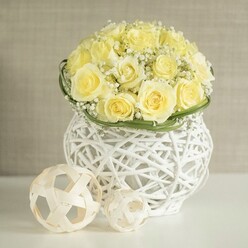 A lush, rounded bouquet of creamy yellow roses dominates the scene, the flowers carefully placed so that each bloom touches the next, creating a luxurious, domed surface that suggests both fullness and softness. The petals, layered in generous spirals, appear almost velvety, catching the light in gentle highlights and shadows that give the arrangement depth. Threaded throughout the roses are delicate tufts of white gypsophila, the baby's breath adding a filigree of tiny blossoms that soften the transitions between the larger flowers and evoke the romantic haze of an early spring morning in Grove Park. Encircling the base of the bouquet is a slim crown of fresh green foliage or flat, woven leaves, which neatly frames the flowers and introduces a crisp, clean line of colour. The arrangement is placed in an eye-catching white vase, its surface formed from interwoven strands that mimic rattan or wicker, rounded in shape with a subtle narrowing at its centre. The open weave allows glimpses of the stems within and lets light pass through, creating intricate patterns on the glossy white tabletop below. Resting to one side are two decorative spheres fashioned from pale wooden or rattan strips, their open, looping geometry playing off the same design language as the vase and adding a sculptural, design-led element to the scene. Behind everything, a softly blurred, neutral-toned wall fades quietly into the background, much like the calm interiors of homes along the quiet streets off Grove Park Road. The composition feels ideal for wedding receptions at local venues, intimate anniversary dinners, or same-day floral deliveries across Grove Park, Hither Green, and the surrounding area, offering a sense of calm elegance, careful craftsmanship, and welcoming warmth.