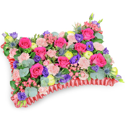 Captured slightly from above on a clean white field, this rectangular floral pillow is a study in colour, texture and quiet craftsmanship familiar to anyone who has ordered sympathy flowers from a neighbourhood shop near Grove Park Station. The top is a pillowy carpet of hot pink roses-some tight and jewel-like, others opening to reveal layered, velvety hearts-softly offset by clusters of pale pink carnations whose frilly edges ripple across the surface. Rich purple lisianthus add cool depth with their cup-shaped blooms, while bright lemon-yellow ranunculus scatter sunny highlights that lift the entire design. Interspersed strategically are steel-blue eryngium thistles, their spiny forms giving a wild, sculptural note that complements the smoother florals. Eucalyptus stems with round, silver-green leaves and varied greenery thread throughout, adding scent and a calming, natural backdrop that allows each flower to read clearly. The pillow's perimeter is defined by a deep red, pleated satin ribbon, ruffled and slightly lustrous, which outlines the rectangular shape and gives a finished, elegant edge. The arrangement conveys warmth and remembrance rather than sombre grief; its palette celebrates life and fond memories. At Grove Park Florist we construct these cushions with dense foam support and careful wiring so they sit respectfully at a service or graveside, and they are often chosen for funerals in the Chinbrook Meadows and Downham areas. From the angle shown, the tribute feels tactile and intimate-a floral embrace offered in difficult moments.