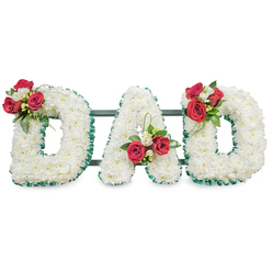 A composed and compassionate sympathy tribute spelling DAD in bold capital letters, each letter densely packed with small, immaculate white chrysanthemums that create a plush, cloud-like surface under soft studio light. The edges of every letter are neatly finished with a ribbed emerald green ribbon that provides a crisp, defining border between the white blooms and the bright background, accentuating the sculpted shapes. On the upper arc of the left D sits a considered cluster of rich red roses and delicate white freesia, softened by lush green foliage; an analogous grouping adorns the centre of the A while the final D repeats the motif, mirroring the composition and reinforcing the sense of unity. Fine loops of bear grass or glossy foliage give a subtle vertical lift within the A, introducing a modern line that contrasts with the traditional chrysanthemum texture. Behind the letters thin dark green or black metal bars are visible, discreetly supporting the scale of the display and ensuring the arrangement travels and stands securely at a funeral or memorial. The scent profile blends the sweetness of freesia and the warmth of roses against the fresh, green undertones of foliage. Crafted with the experience of local florists who work daily for services around Grove Park station, Baring Road and Marvels Lane, this design is suited to chapel services or graveside farewells and can be prepared for same-day attention across Grove Park and nearby Lee. It conveys solemnity, deep affection and a personalised farewell to a beloved father or paternal figure.