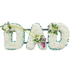 A three-dimensional floral display spelling out DAD is the sole focus of this photograph, the letters standing tall on a subtle dark frame that disappears into a white studio backdrop. Each generous block letter is completely covered with immaculate white chrysanthemum heads, meticulously placed so that every petal touches its neighbour, creating a soft yet structured blanket of blooms. The overall effect is one of gentle calm and purity, a fitting choice for a final tribute. Around each letter runs a rich, emerald green satin ribbon, folded into small ruffles that follow the contours and lend crisp definition, echoing the lush greenery and riverside walks that characterise Grove Park. The first D carries a small corsage of white freesias and fresh foliage at its upper left, their slender buds and open trumpets adding lightness and a touch of movement. From this point, two delicate green stems sweep diagonally across the arrangement, leading the eye toward the central A, where an elegant focal spray has been arranged. Here, two deep purple calla lilies, with velvety, trumpet-shaped blooms, are nestled amongst white freesia flowers and vivid green leaves, bringing a subtle but striking contrast to the predominantly white design. The last D is finished with a smaller, harmonious cluster of freesias and foliage on its top right curve, mirroring the embellishment on the first letter without overpowering the simplicity of the tribute. The design conveys dignity, tenderness, and gratitude, the sort of carefully composed arrangement that might be delivered by a local Grove Park florist for a funeral or memorial gathering to honour a devoted dad.