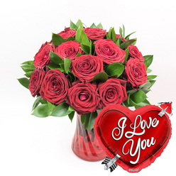 This image captures the essence of a romantic gift you might send to someone special in Grove Park, with every detail crafted to speak of love and thoughtfulness. At its core is a lush bouquet of deep red roses, gathered in a rounded, generous cluster. Around a dozen or more blooms are visible, their velvety petals unfurling in rich layers of crimson. Some roses are fully open, revealing their intricate centres, while others remain slightly cupped, creating a dynamic mix of shapes and textures that feels both natural and expertly arranged. Nestled among the blooms is a collar of fresh, vibrant green foliage, the leaves smooth and gently arching. This greenery frames the roses and adds a crisp contrast that makes the red tones glow more vividly, reminiscent of the vibrant colours seen in local gardens near Grove Park and Gunnersbury. The stems of the bouquet are gathered into a clear, ruby-red glass vase with subtle fluted lines running vertically along its sides. These delicate ridges catch the light, creating soft reflections and giving the vase a sense of movement and refinement. The red tint of the glass echoes the colour of the petals, unifying the overall palette into a warm, romantic harmony. Resting in the lower right foreground, and slightly overlapping the vase, is a gleaming heart-shaped foil balloon in bright, metallic red. Its surface is smooth and glossy, with highlights and reflections that give it a playful, festive feel. Across the centre of the heart, in flowing white script, the words I Love You are clearly written. A silver arrow, evoking Cupid's arrow of legend, appears to pierce the heart diagonally from bottom left to top right. Against a pristine white background, these elements combine to form a striking image of devotion, perfect for anniversaries, Valentine's surprises, or any occasion when you want a floral gift to speak clearly of your feelings in Grove Park.
