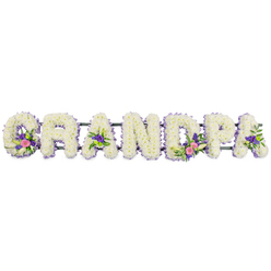 The image presents a personalised funeral flower arrangement in the form of the word GRANDPA, crafted with the care and precision you might expect from a specialist florist working for a family in Grove Park. Seven individual capital letters are arranged in a straight line, supported by a slim, dark green frame that is only just visible behind the blooms. Each letter is entirely packed with small, white chrysanthemums, their layered petals pressed closely together so the surface looks plush and soft, almost like freshly fallen snow. Around the outer edge of every letter, a fine trim of lilac and purple flowers adds a delicate border, defining the contours and lending a gentle, soothing colour contrast that remains respectful and calm. Several letters are decorated with extra accent sprays: on the 'G' and the first 'A', sprays of bright pink and rich fuchsia flowers nestle among glossy green foliage and slender white lily-like flowers. The 'D' and another 'A' also carry these colour accents, pairing bright pinks and purples with fresh greens that lift the predominantly white design without overwhelming it. The entire word is photographed against a perfectly plain white background, under even lighting that brings out the crisp whites, cool purples, and warm pinks. It is easy to imagine this tribute resting at a funeral in one of the churches near Grove Park station or being carried to a graveside at nearby cemeteries, where it would stand as a bold yet gentle statement of love. The arrangement exudes quiet dignity, nostalgia, and gratitude for a grandfather's life, offering a floral embrace when words feel too small.
