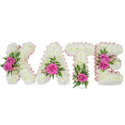 Crafted by experienced hands in the Grove Park area, this personalised floral tribute spells the name KATE in four substantial, freestanding letters that appear almost to glow against a clean white backdrop. Each letter is composed of hundreds of compact, pristine white chrysanthemums pressed close together to form a continuous, cloud-like face, and the inner edges are softly defined with a pale pink satin ribbon that lends a refined, feminine outline. Hot pink roses act as intentional visual anchors: two open roses grace the left side of the K, the A holds a single, fully opened rose near its base surrounded by small pale-pink buds, the T is balanced by one vivid pink rose at its lower centre framed with ferny foliage, and the E is brightened by a pair of roses on its right edge accompanied by variegated green leaves. Additional greenery - hints of eucalyptus-like ruscus and slender spiral grass - is threaded through the white blooms to add depth and natural movement, while the overall finish is neat and three-dimensional, with soft shadows that suggest the letters sit slightly raised from their surface. There is a faint perfume of roses that complements the matte white chrysanthemum, a restrained and comforting scent appropriate for a funeral or remembrance display. Available for local delivery around Grove Park station, Downham and Hither Green, Grove Park Florist can recreate this tribute to reflect a loved one's personality and provide a clear, heartfelt expression at a memorial service.