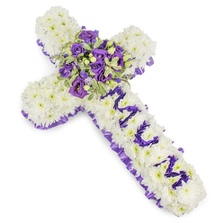 This large Christian cross tribute presents a dense carpet of pristine white chrysanthemums, each flower placed so closely it creates a smooth, cloud-like surface that suggests purity and gentle reverence. A ruffled purple satin ribbon traces the entire outer edge of the cross, the deep hue and subtle sheen giving the piece a formal frame that complements the softness of the petals. The vertical arm carries a personal message: MUM, spelled clearly in rich purple flower heads - likely small lisianthus or spray chrysanthemums - their velvet tones standing out against the white backdrop. At the intersection of the arms a generous central spray becomes the visual heart, composed of deep violet and lavender roses with their tight, layered petals, intertwined with lighter purple lisianthus and eustoma that add ruffled texture, while cream-edged variegated foliage and tiny green berries tuck into the cluster to provide contrast and depth. The bouquet sits on a stark white background under gentle, even lighting so every petal and leaf reads with clarity. As crafted for families across Grove Park, Lee and Hither Green, this arrangement carries the quiet dignity appropriate for funeral chapels, wakes, or cemetery services; Grove Park Florist prepares each piece with the same careful hand we would use for deliveries to Baring Road or Grove Park station area. Tactile, fragrant, and thoughtfully balanced in tone - from the cool whites to the solemn purples - the tribute expresses enduring love and measured sorrow in equal parts.