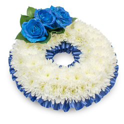 Seen from just above, on a crisp white background that emphasizes form and colour, this traditional funeral wreath is composed with quiet precision. A generous, slightly domed ring of white chrysanthemums makes up the main body; each blossom displays delicate, plume-like petals that create a soft, tactile surface and are warmed by tiny, creamy-yellow eyes in their centres. Nestled to the upper left are three intense blue roses - their petals rich and velvety, opening into tight spirals that draw the eye and introduce a dignified, solemn note. Dark green foliage sits directly beneath the blue grouping, providing a fresh, living counterpoint and allowing the blue to read as both distinct and integrated. Framing the chrysanthemum ring is a pleated satin ribbon in matching cobalt blue, folded and ruffled along the inner and outer rims so the arrangement reads as a carefully finished, bordered piece; the ribbon's sheen lends a formal, respectful shimmer without overpowering the flowers. The soft, diffuse lighting captures texture and depth: petals, leaves, and satin reflect small highlights, while a gentle shadow beneath the wreath gives it presence and dimensionality. As a local florist who often prepares tributes for services near Baring Road and around Grove Park station, I picture this wreath laid beside a floral table or standing at a remembrance service, its colours chosen to console and honour. The overall impression is composed, serene, and fitting for moments of farewell or quiet commemoration.