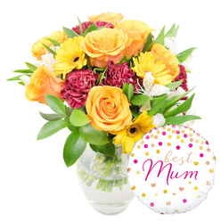 This image presents a thoughtfully composed Mother's Day gift set that reads as both cheerful and expertly arranged. Dominating the left of the frame are sumptuous orange and yellow roses, their petals unfolding in layered spirals that catch soft studio light and suggest a warm, citrus-tinged scent. Bright yellow gerbera daisies intersperse among the roses like small suns, their dark brown centres giving the arrangement visual rhythm, while rich maroon carnations contribute dense, velvety texture and romantic weight. Slender white alstroemeria threads delicately between the bolder blooms, its fine speckling and narrow petals introducing an elegant counterpoint. A lively assortment of deep green foliage-glossy leaves and feathery sprigs-wraps the composition, offering both structure and a fresh, earthy contrast. The stems rest in a clear glass vase filled with water, the transparency underscoring the bouquet's newly-arranged quality and careful preparation. Nestled to the lower right, partly overlapping the vase, a round white foil balloon decorated with gold and hot-pink polka dots and tiny hearts proclaims best Mum in flowing hot-pink script, giving the image an unmistakable celebratory focus. As a local florist serving Grove Park, Lee and Hither Green, Grove Park Florist often pairs blooms and balloons like this for mothers who live near Chinbrook Meadows or need a thoughtful doorstep surprise; the photo conveys the tactile softness and joyful intention behind such a gift without losing the quiet craftsmanship of the arrangement.