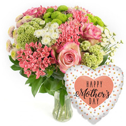 This image presents a beautifully styled Mother's Day bouquet paired with a festive balloon, photographed in soft, even light against a pure white backdrop so that nothing competes with the colours of the flowers. The bouquet is arranged in a simple clear glass vase, its transparency revealing the carefully gathered stems and clear water within, a small detail that speaks of freshness and professional care. Above the vase, the flowers form a rounded, slightly domed shape. Several large pink roses occupy the top and centre, their petals unfolding in gentle spirals, edges kissed with a slightly darker pink, suggesting tenderness and affection. Intermingled with the roses are clusters of small coral-pink flowers similar to kalanchoe, their numerous tiny petals creating a dense, textured surface that draws the eye closer. Lime-green button chrysanthemums punctuate the arrangement, each one a neat, circular burst of colour with a delicately frilled finish, adding energy and a contemporary feel. Between these brighter tones, white spray chrysanthemums with daisy-like faces offer a soft, airy lightness, balancing the composition and adding a sense of calm brightness that reflects the clear sky often glimpsed over Grove Park. Lush dark green foliage fills the lower sections and peeks through between blooms, outlining the bouquet with natural depth. To the right of the flowers, a heart-shaped foil balloon leans slightly towards the arrangement. Its pale background is scattered with shimmering gold and soft peach confetti-style dots, while at the centre a solid peach heart carries the inscription HAPPY Mother's DAY in elegant black script and bold lettering. Together, flowers and balloon evoke a warm, heartfelt celebration, as if ready to be delivered from Grove Park Florist to a mother relaxing at home near Chiswick or the tree-lined streets by the station.