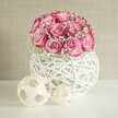 A softly lit indoor scene shows a lush, rounded bouquet of pink roses and white baby's breath resting in a modern, woven white vase, styled as if on a calm morning in a Grove Park living room. Around fifteen to twenty roses form a dome of colour, each bloom opening generously so the petals can be seen graduating from pale blush at the centre to a deeper, almost fuchsia pink at the edges. The roses look velvety and full, their spiralling petals creating a sense of movement, as though the bouquet has just been turned on the table for a final check. Between the roses, tiny stars of white Gypsophila float like clouds, softening the pinks and adding that familiar, delicate florist's scent you'd notice when stepping into a local Grove Park flower shop. The flowers are gathered into a spherical white vase crafted from criss-crossing strands that resemble painted wicker or rattan, airy and open so that light passes through to cast gentle shadows on the smooth white surface below. To the left of the vase sit two decorative spheres made from light, natural fibres in a sandy beige tone, their open, geometric weave echoing the texture of the vase and bringing a touch of Scandinavian-style simplicity to the composition. Behind, a warm beige wall, subtly textured, provides a quiet backdrop that allows the pink roses to glow. The overall mood is peaceful and romantic, ideal for a coffee table in a Grove Park flat, or as a thoughtful anniversary or Mother's Day gift that feels both contemporary and heartfelt.