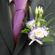 A compact study in contrast and calm, this boutonnière combines delicate craftsmanship with the fresh simplicity of garden blooms. At its centre sits a white lisianthus, the edges of each petal kissed by a striking purple that throws soft shadows and definition across the blossom; nearby a white dianthus with lively purple ruffles adds a more structured, textural note that complements the lisianthus's softness. Slender lisianthus buds and a single upright stem of indigo veronica introduce vertical rhythm, while feathery ornamental grasses and dark green foliage create a natural cradle around the flowers. Scattered clusters of gypsophila lend breathy lightness, like small clouds of white, and the whole is gathered with a crisp white ribbon whose tidy knot signals careful finishing. Laid against the lapel of a dark suit and paired with a magenta silk tie, the boutonnière reads as refined but personable-perfect for a groom or honoured guest at celebrations across Grove Park and neighbouring areas. As a local touch, our team at Grove Park Florist often pairs this look with weddings held near Grove Park station and community halls nearby, where the quiet scent and fresh textures mingle with the afternoon air. The arrangement feels composed yet alive: the purple edging gives drama, the buds promise growth, and the greenery keeps the overall impression grounded and sincere throughout a long, memory-filled day.