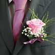 This boutonniere, styled with the calm assurance of an experienced Grove Park florist, features a single blush pink rose resting against the dark structure of a charcoal suit. The bloom itself feels like the visual heartbeat of the piece: velvety petals in a soft, romantic pink unfurl from a tight centre, forming gentle curves that reflect the light with a subtle, almost satiny sheen. Encircling the rose, dainty stems of white gypsophila scatter tiny round blossoms, giving the arrangement a delicate, airy halo. These little flowers add texture that contrasts beautifully with the smooth rose petals and the crisp weave of the suit fabric. Threaded among them, slender blades and curls of fresh green foliage bring a hint of lively movement, a reminder of gardens and riverside walks near Grove Park and the Thames. At the base of the boutonniere, the stems are firmly bound in a soft lilac-pink ribbon, its polished surface catching the light just enough to underscore the care taken in its tying. The ribbon's colour echoes the rose's blush, uniting the elements into one harmonious composition. The lighting in the image is gentle and flattering, enhancing the depth of the pink tones and the brightness of the white baby's breath without overpowering the natural character of the flowers. It is easy to imagine this boutonniere worn at a wedding in one of Grove Park's churches, at a reception in a tucked-away garden venue, or even at a formal event across nearby Chiswick. The overall impression is of quiet sophistication and understated romance, a floral detail designed to be both seen and fondly remembered.