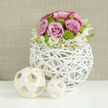 On a crisp white table, polished to reflect just a hint of light, an elegant floral centerpiece captures the mood of a modern Grove Park wedding or engagement celebration. The flowers are gathered in a globe-shaped vase woven from slender, white-painted branches, each piece crossing and looping to create an open lattice that is both rustic and architectural, reminiscent of the tree canopies in nearby Chiswick or along Strand-on-the-Green. Rising from this sculptural base is a perfect dome of blooms: pastel pink roses, some edged with a darker rose hue, whose velvety petals curl open in generous layers, forming a soft, romantic canopy. Threaded among them are clusters of vivid lime-green button chrysanthemums, their small, rounded flower heads adding a playful burst of colour and textural contrast that keeps the design contemporary. At the base of the arrangement, delicate white flowers with tiny yellow centres spill gently over the rim, disguising the mechanics and melting into the woven branches below. To one side, two decorative balls made from thin, pale wooden strips sit on the tabletop, echoing the spherical shape and airy pattern of the vase and tying the whole composition together. Behind, a warm neutral background with a subtle, linen-like texture is softly out of focus, allowing the eye to linger on each petal and twig. The effect is one of quiet sophistication and joyful freshness, perfectly suited to a Grove Park reception table, registry office celebration, or intimate family gathering where guests are welcomed with thoughtful, beautifully crafted flowers.